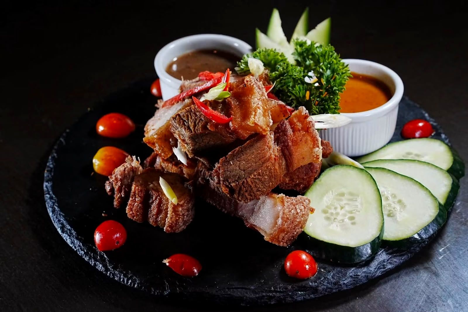 Crispy pork on a dark slate plate with two dipping sauces, cucumber slices and cherry tomatoes