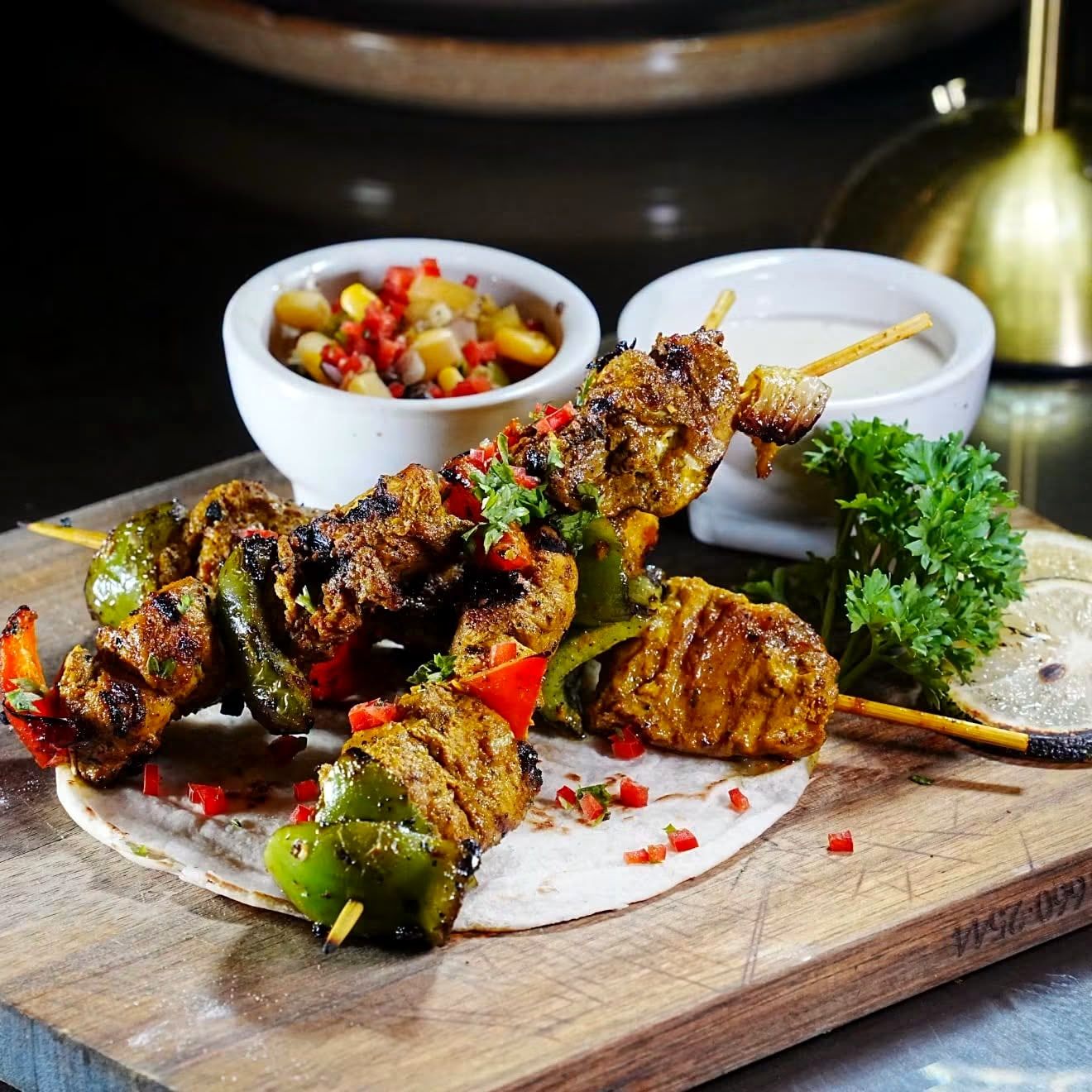 Grilled chicken skewers with peppers on a flatbread, served on a wooden board with two small bowls on the side