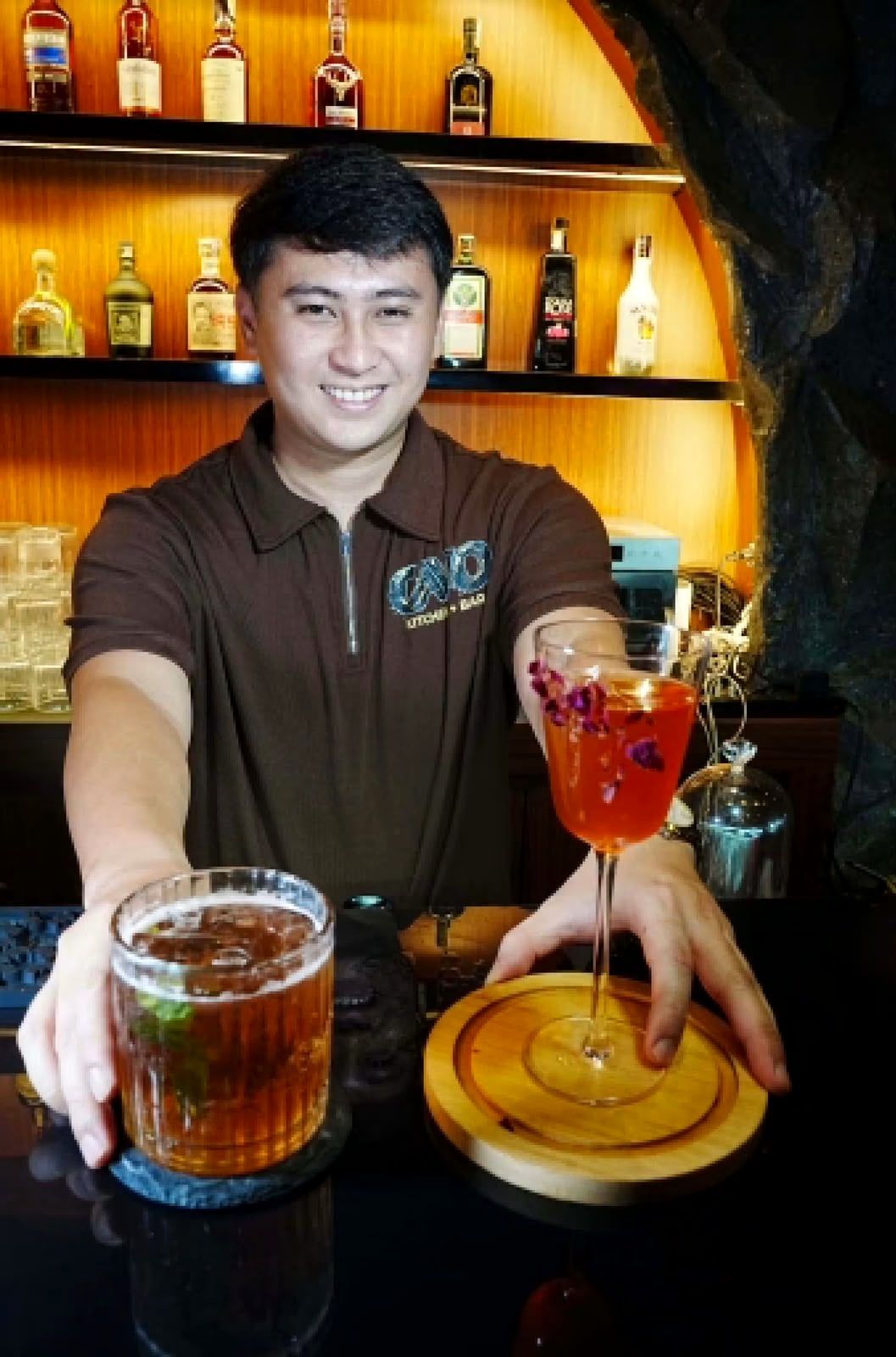 A bartender in a Cavo polo shirt presenting two cocktails in front of the lit bar shelves