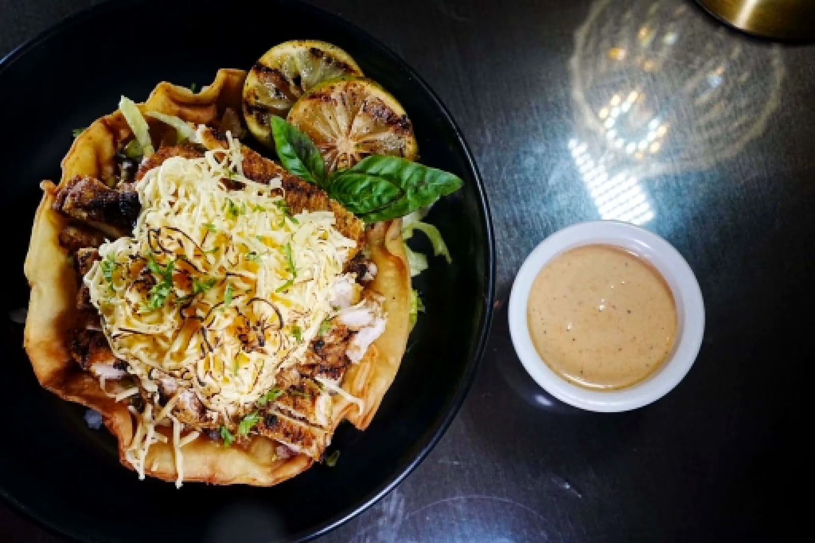 A filled crisp tortilla bowl topped with shredded cheese, seen from above beside a small bowl of sauce