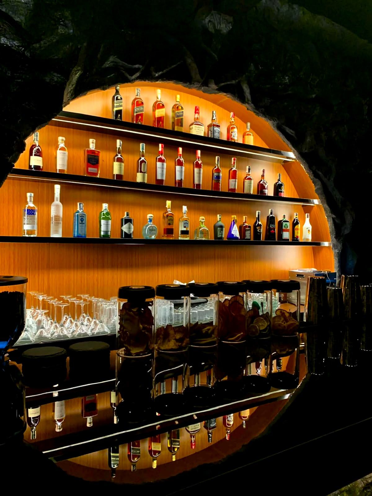 Bottles on lit amber shelves inside the rock arch, with glasses lined up on the counter below