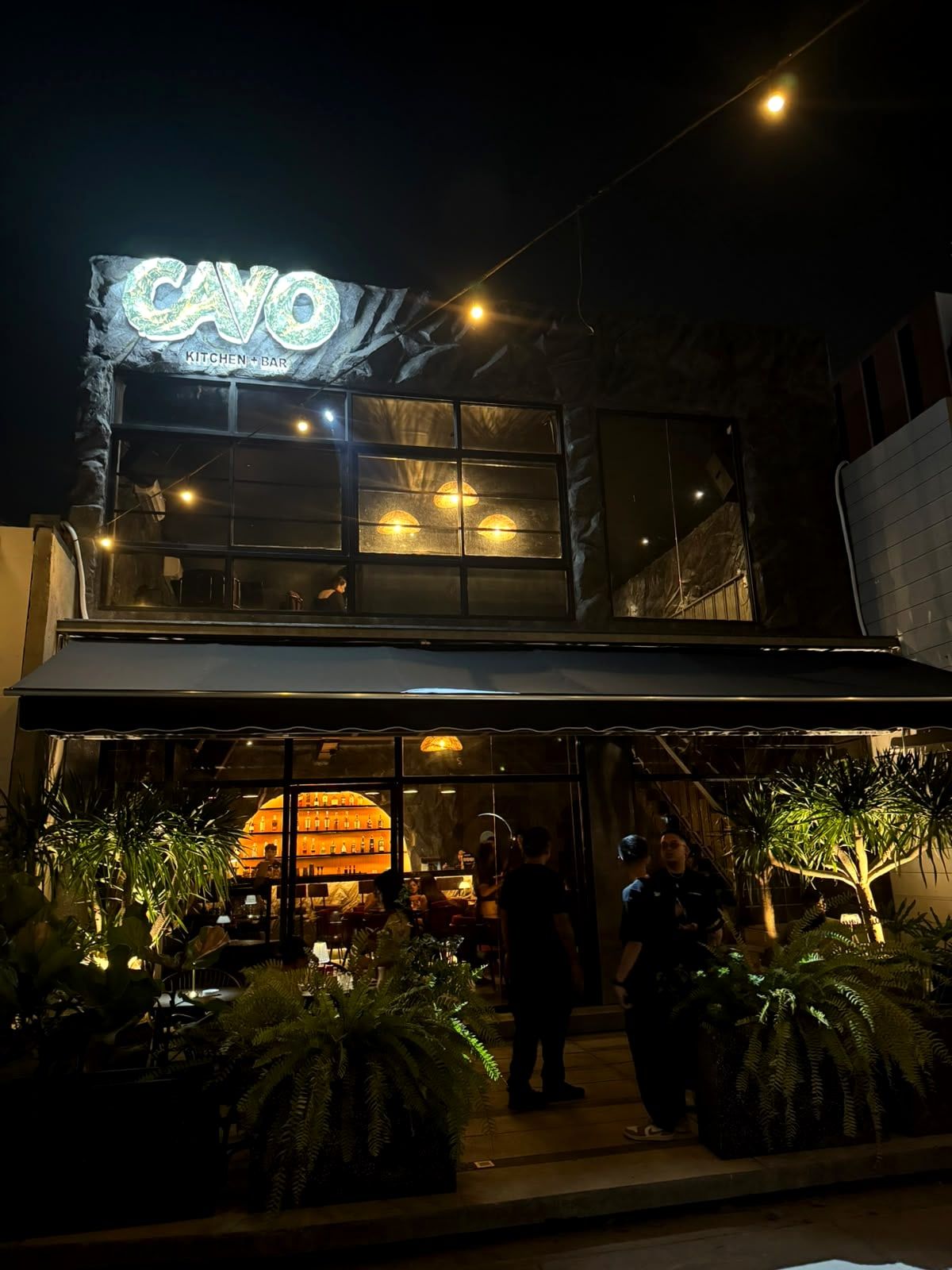 The front of Cavo at night: the lit CAVO Kitchen + Bar sign above tall windows, with guests at the entrance between green plants