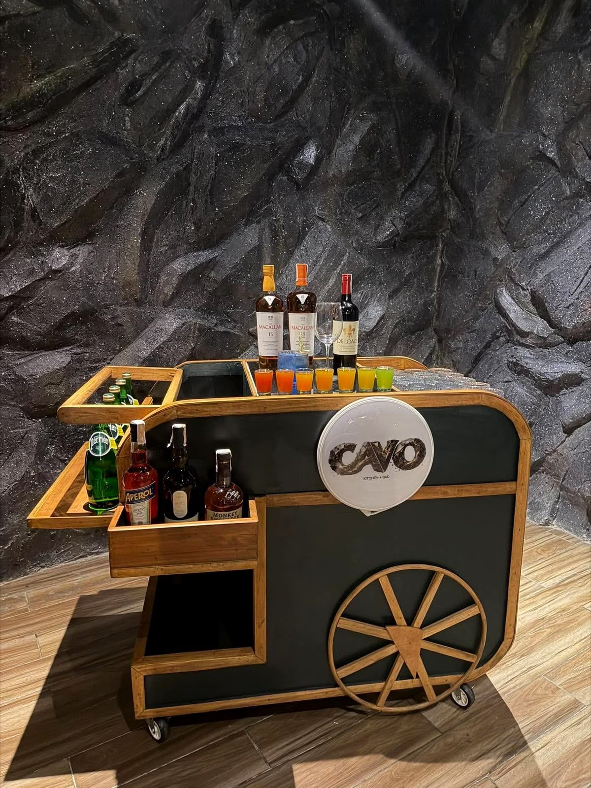 The Cavo mobile bar: a dark green cart with wooden wheels, the CAVO logo, bottles and a row of shots
