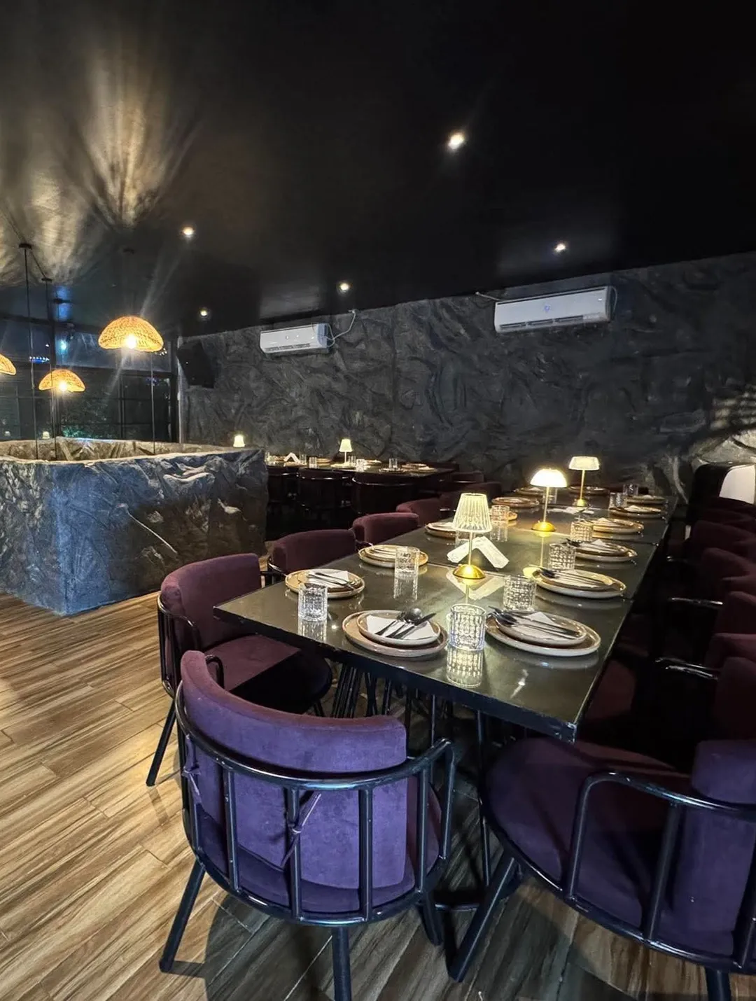 The upper floor set for dinner: a long table with purple chairs and table lamps against the rock wall