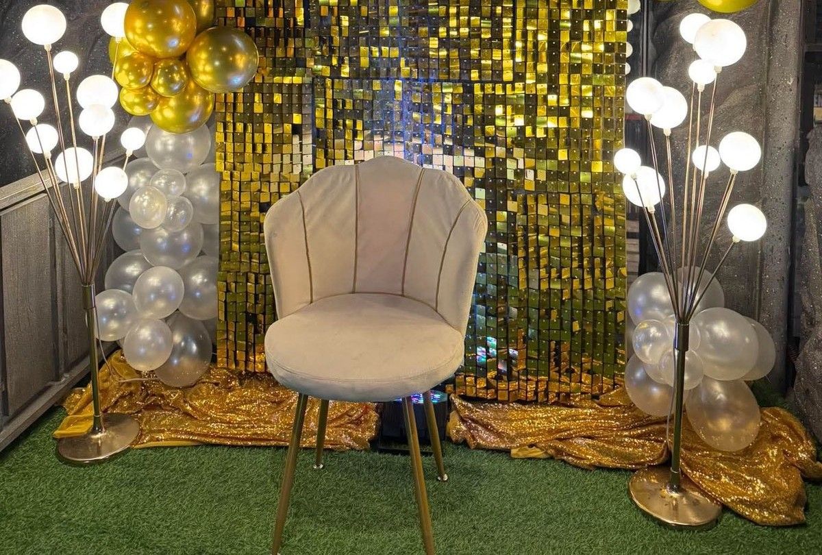 A gold sequin backdrop with balloons, warm lights and a white shell chair, set for a birthday
