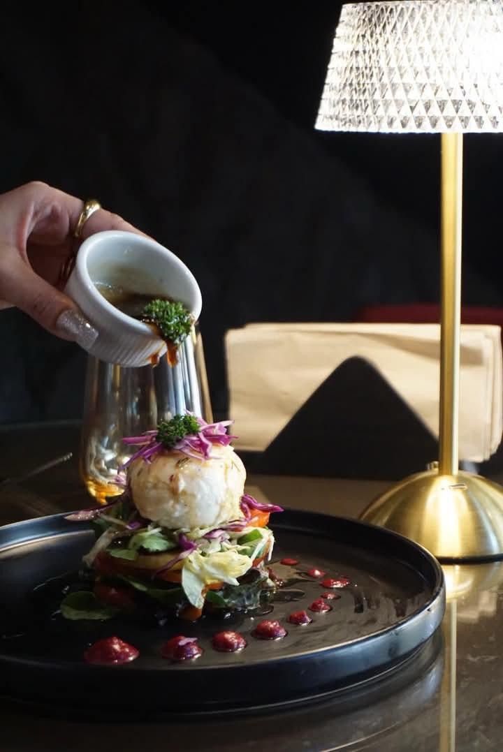 Sauce poured from a small jug over a stacked plated dish, under a brass table lamp