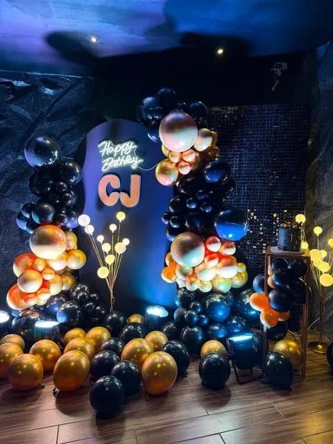 A birthday balloon garden in black, orange and gold with a neon sign